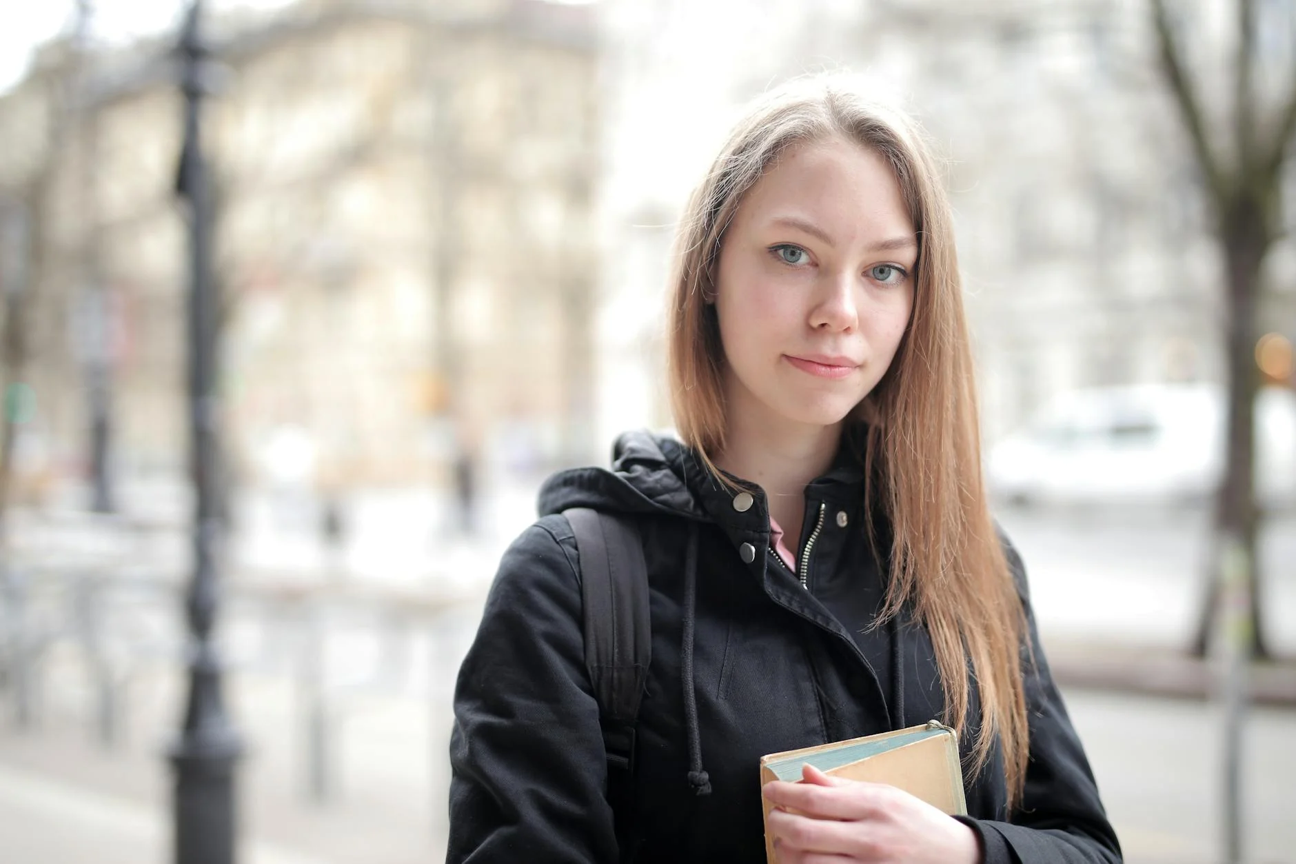 Portrait of a young woman smiling outdoors holding a book on a city street. Casual and contemporary setting.