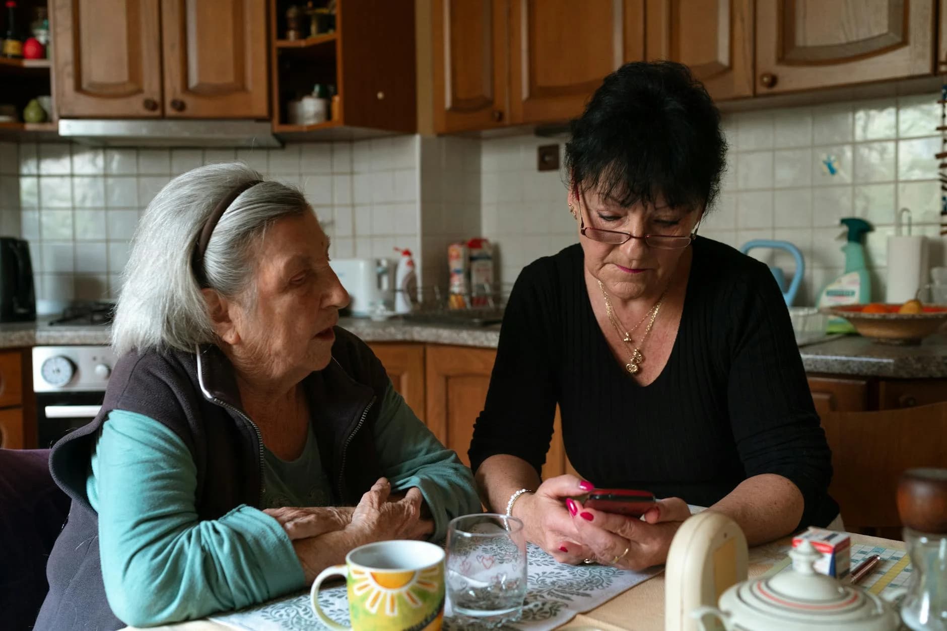 A senior woman and a caregiver share a moment in a warm, home kitchen environment in Prague.