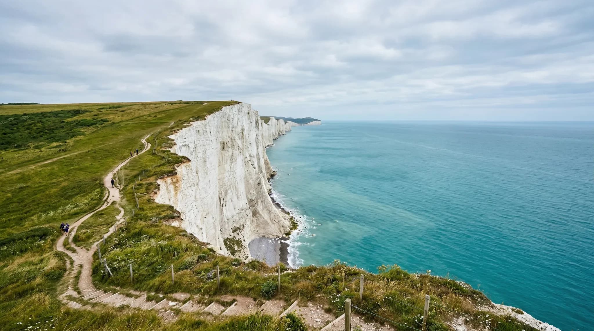 A wide-angle photorealistic landscape shot of towering white chalk cliffs overlooking a calm turquoise sea under a soft, overcast northern European sky. The cliff edge is topped with patches of green