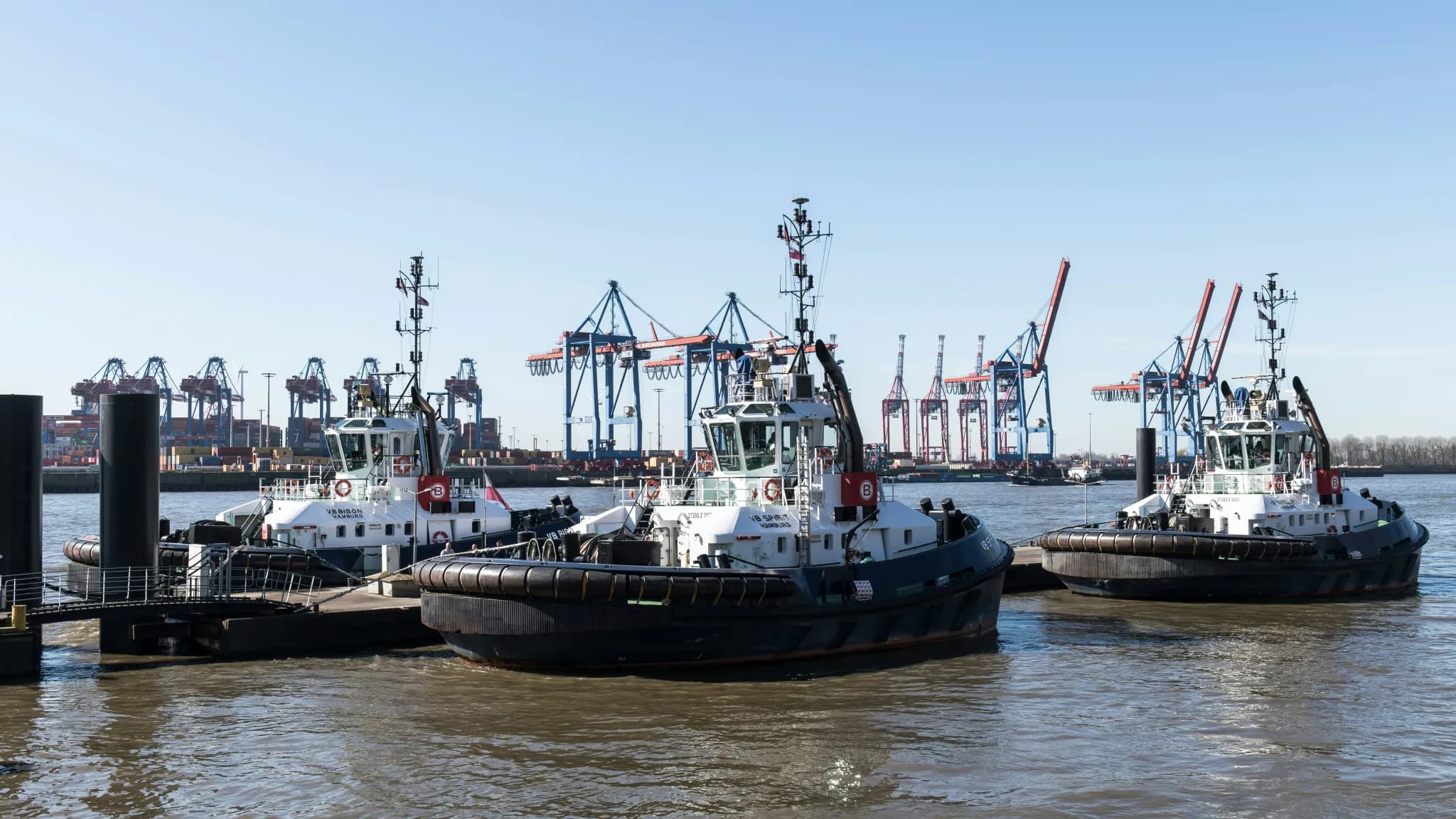Three tugboats docked at a harbor with cranes in background