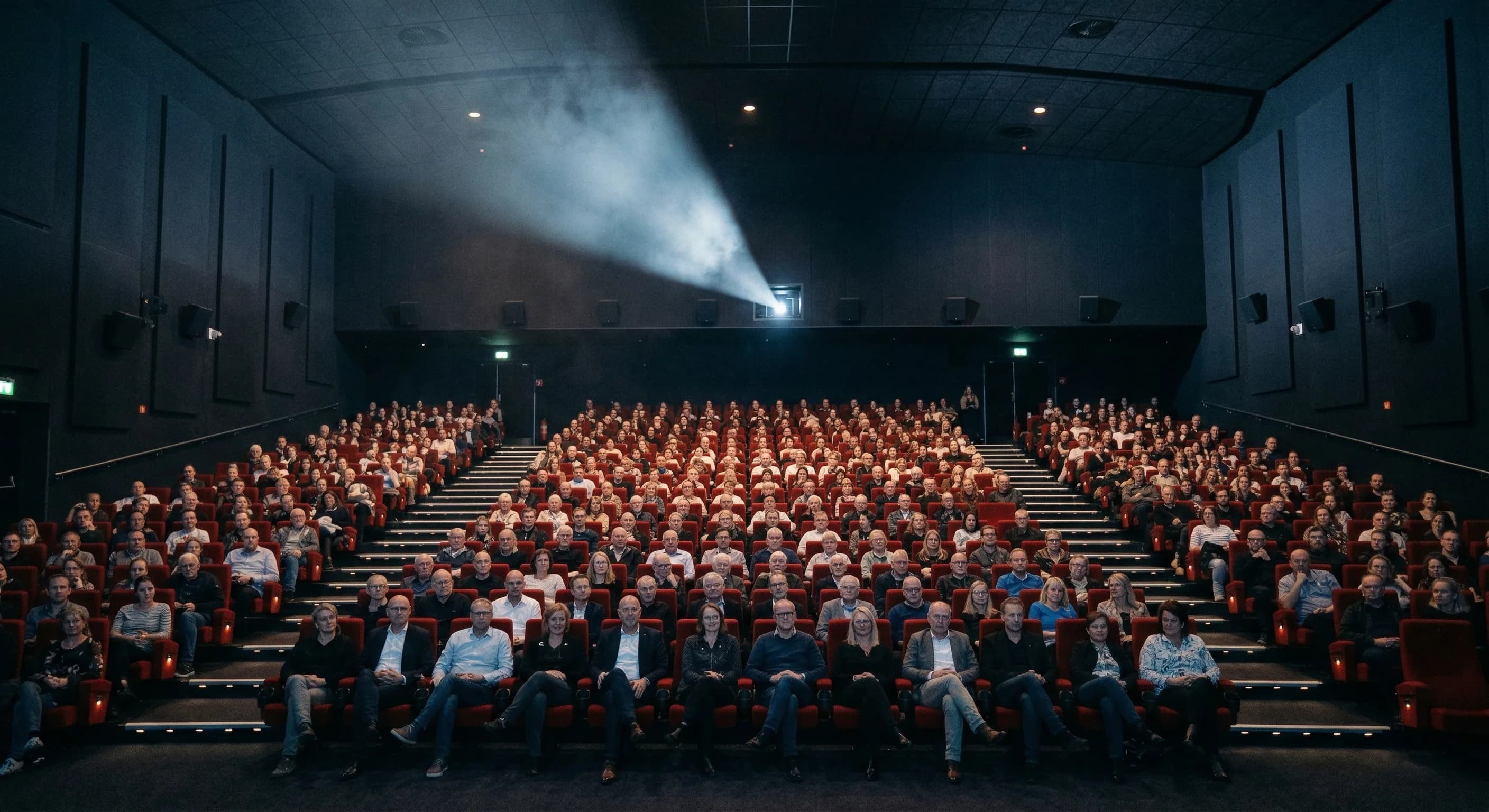 A wide-angle photorealistic shot from the back of a modern cinema auditorium in Northern Europe. The rows of deep red velvet seats are filled with people of various ages, their silhouettes softly illu