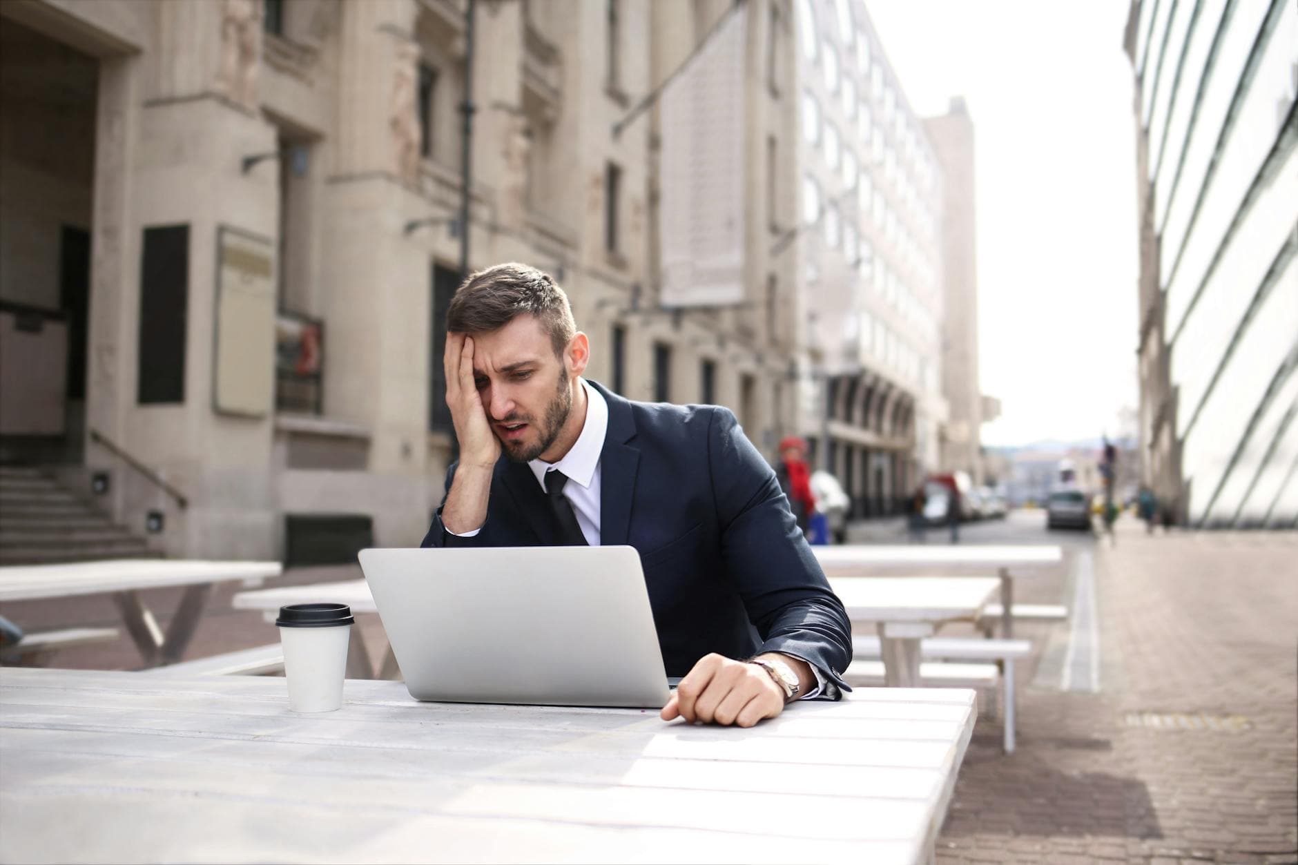 A businessman in a black suit looks stressed while working outdoors on a laptop with coffee.