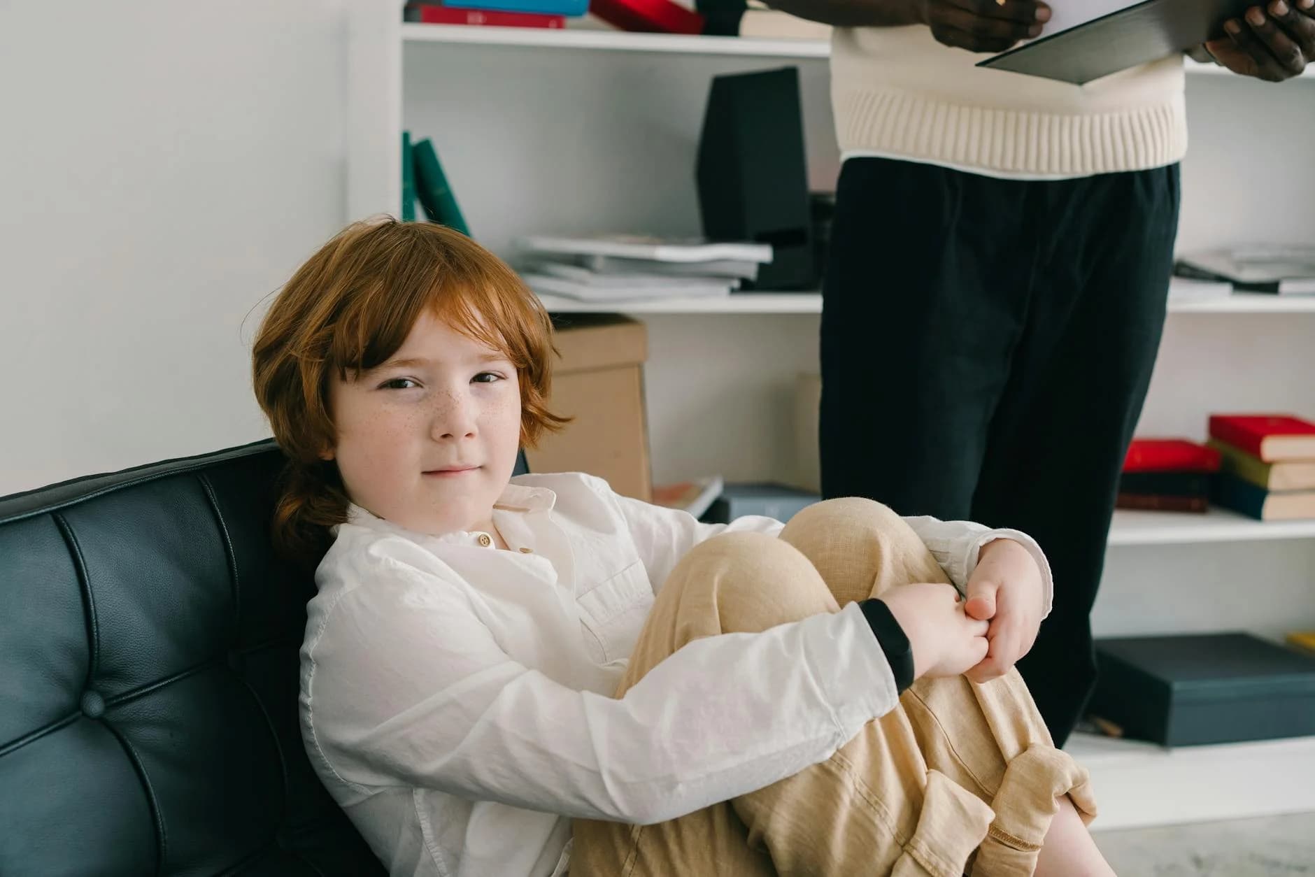A young boy with ginger hair and freckles sits comfortably on a black leather chair, looking calmly at the camera in what appears to be a therapist's office. In the background, a person stands holding a clipboard, contributing to a professional yet supportive atmosphere.