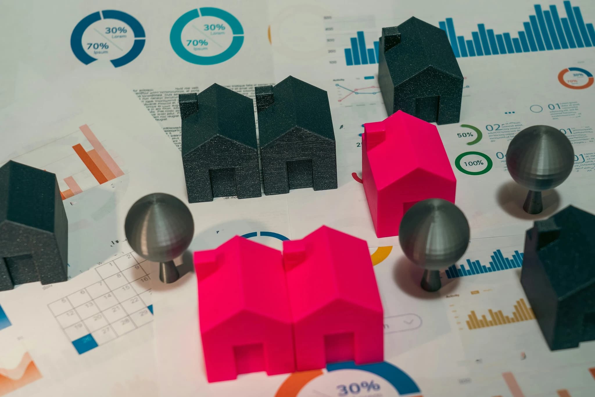 The image features a collection of 3D-printed black and bright pink house models and small spherical "trees" arranged across a surface covered in financial charts, graphs, and a calendar. The composition creates a metaphorical scene of real estate planning or market analysis, blending physical models with analytical data to suggest a mood of strategic property development.