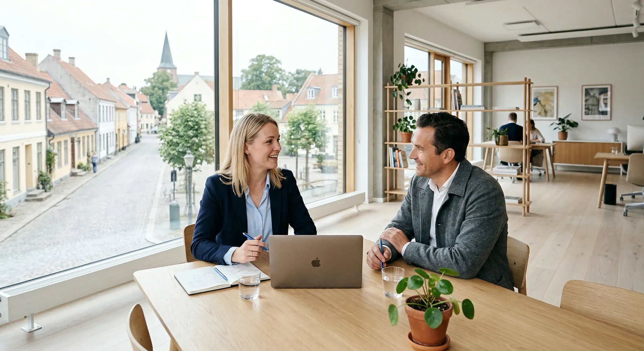 A modern, bright office space with clean Scandinavian interior design. A consultant and a visitor are engaged in a professional conversation at a light oak table. Large windows in the background let i