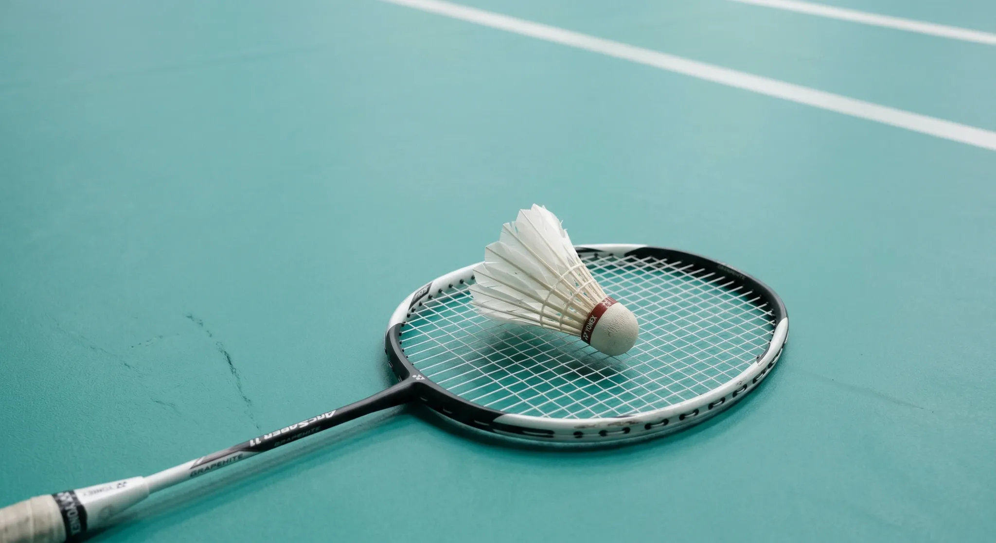 A minimalist composition featuring a single white feathered shuttlecock resting on the strings of a modern badminton racket. The racket lies on a clean, matte teal-colored sports court floor. The ligh