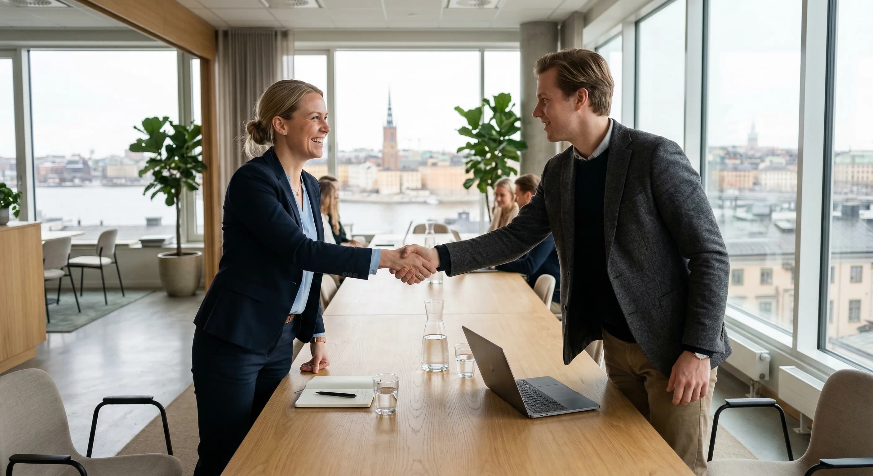 A photorealistic wide-angle shot of a professional handshake occurring over a light oak conference table in a modern Scandinavian office. The room is filled with soft, natural light from large floor-t