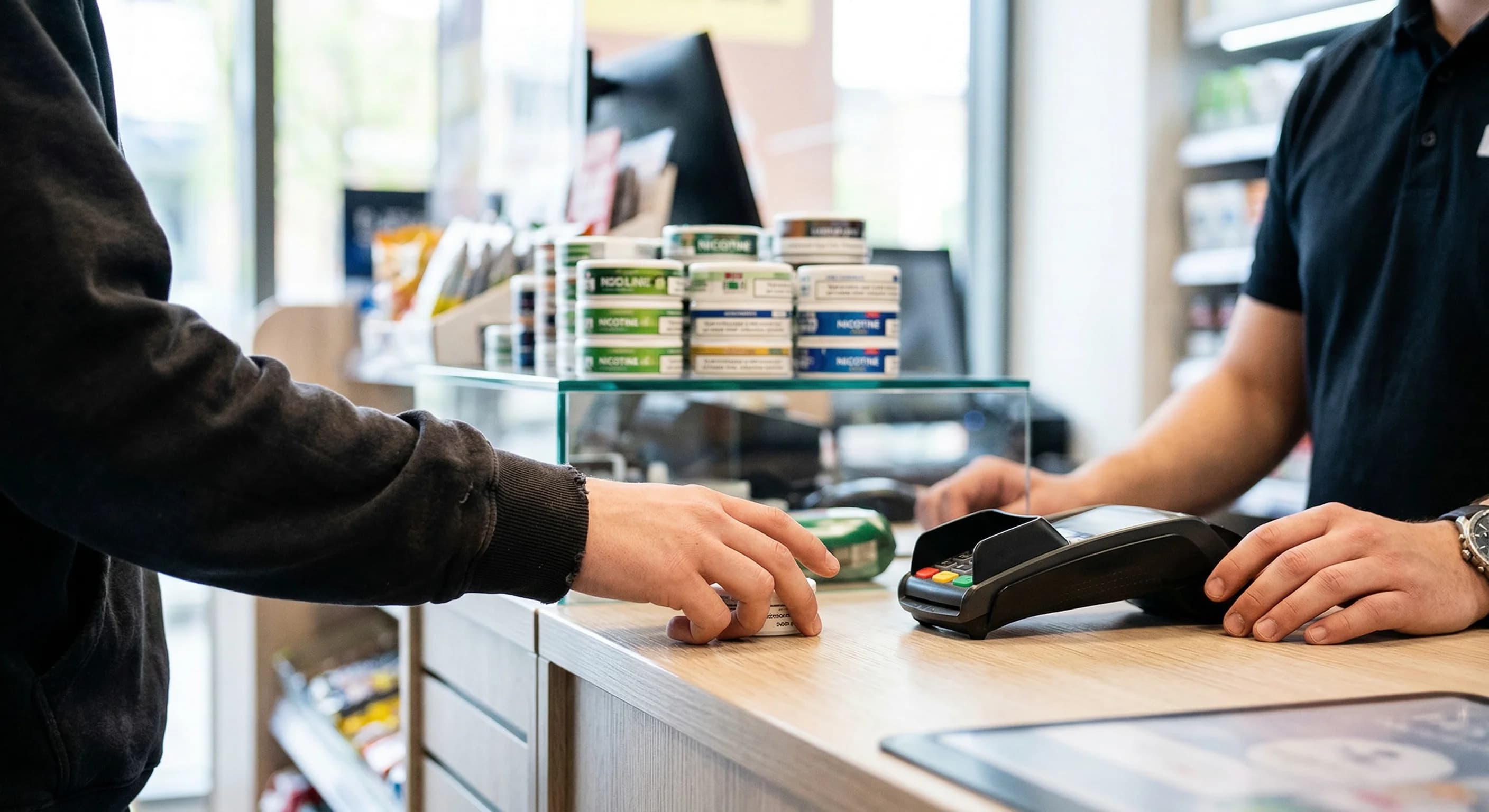 A close-up, shallow depth of field shot of a modern retail counter in a kiosk. Several small round tins of nicotine pouches are stacked on a glass shelf in the background, slightly out of focus. In th