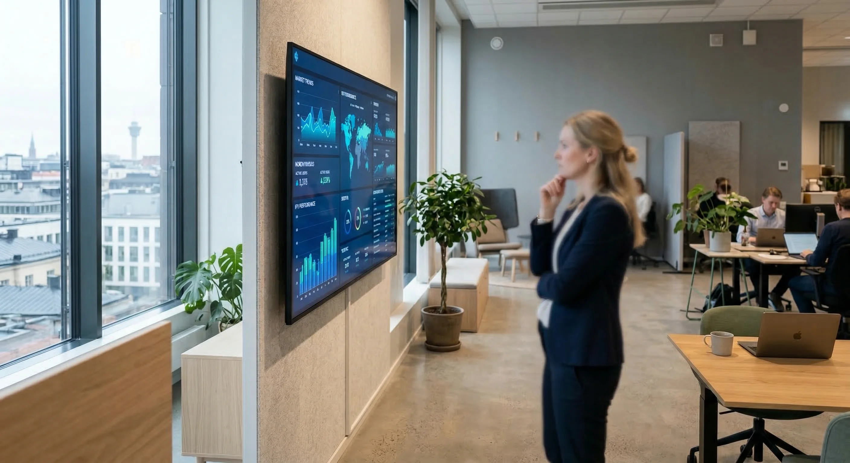 A clean, modern office interior in Northern Europe featuring a large wall-mounted digital dashboard with bright blue and teal data visualizations. A professional person is seen from the side, partiall