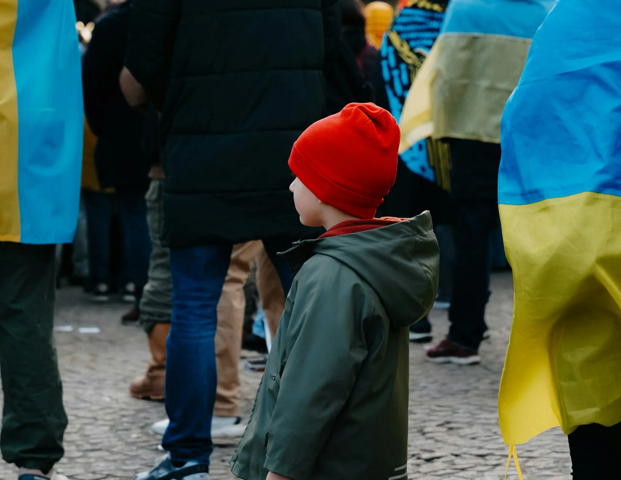 A young child wearing a bright red beanie and a green jacket stands among a crowd draped in Ukrainian flags during a public demonstration. The scene conveys a somber, reflective mood as the child looks off into the distance amidst the sea of blue and yellow colors.