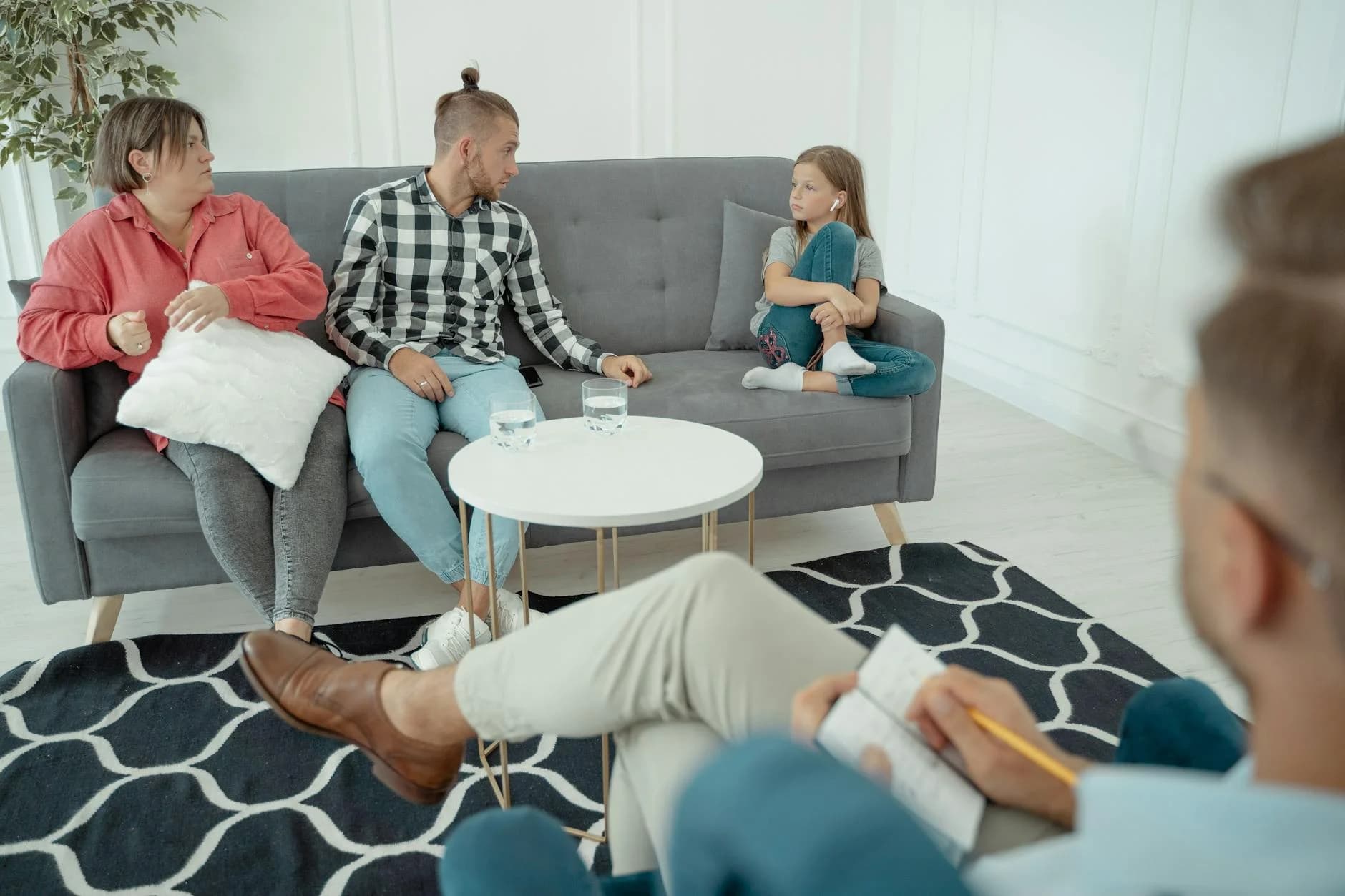 A family sits on a gray sofa during a therapy session, with the father looking toward his daughter, who is wearing earbuds and appears disengaged. The setting is a bright, minimalist office, where the therapist in the foreground takes notes, creating an atmosphere of professional guidance amid palpable interpersonal tension.