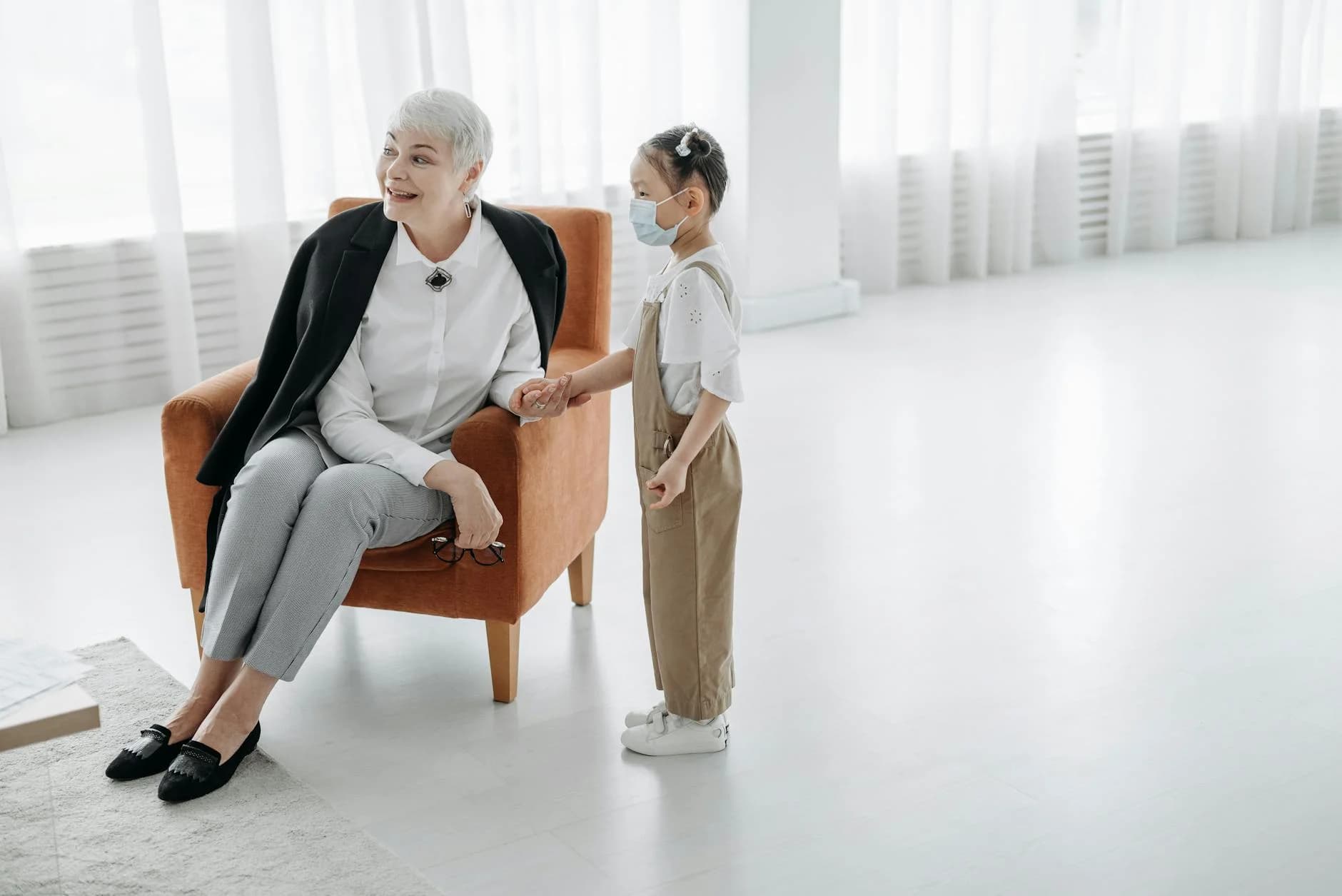 An elderly woman in business attire sits in an armchair, smiling and holding hands with a young child wearing a face mask in a bright, minimalist room. The scene evokes a gentle and supportive atmosphere, suggesting a professional yet caring introduction or meeting.
