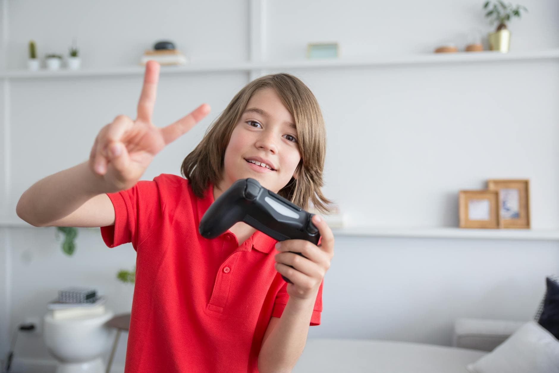 Young boy in a red polo shirt playing video games and flashing a peace sign indoors.