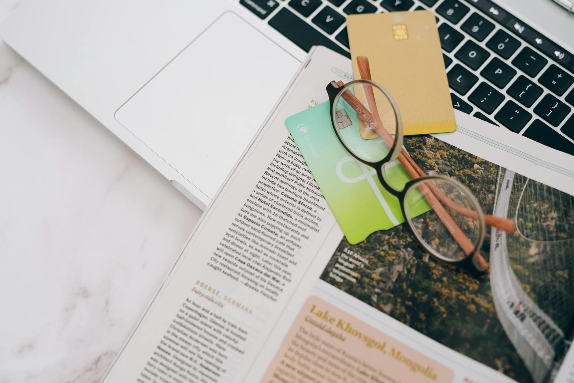 Eyeglasses resting on a magazine beside a laptop and credit cards, indoors.
