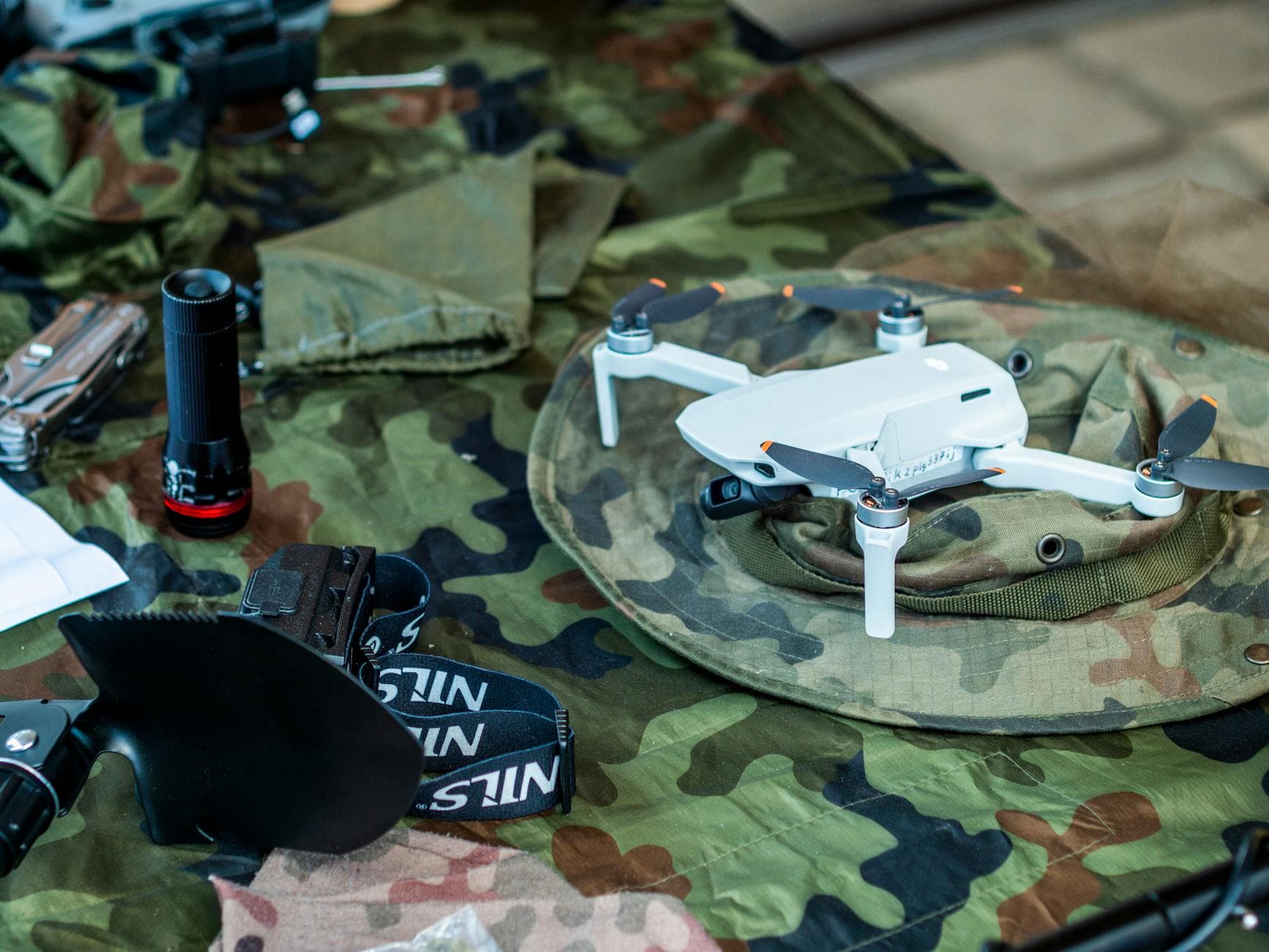Compact white drone on camouflage gear, surrounded by flashlight and tools.