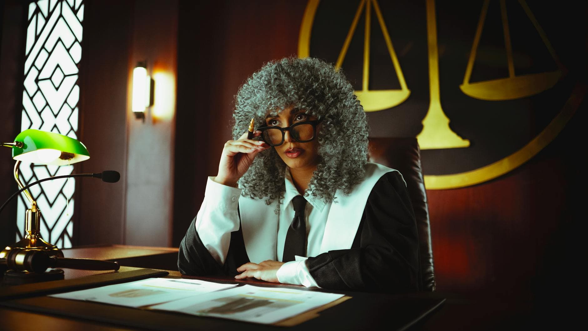 Judge in traditional attire sits at a courtroom desk with a serious expression.