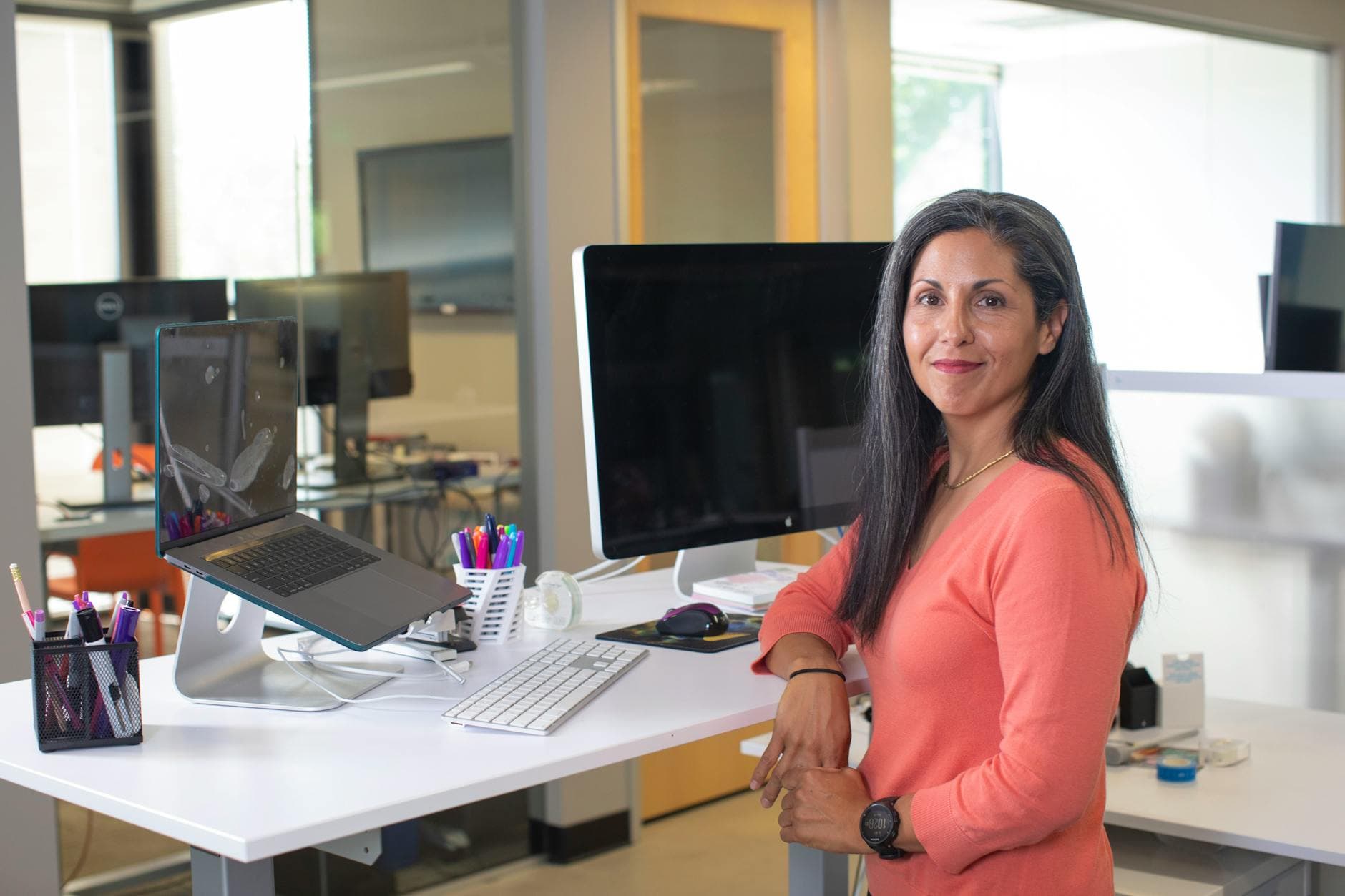 A woman in a pink top smiles confidently in a modern office with computers and desk accessories.