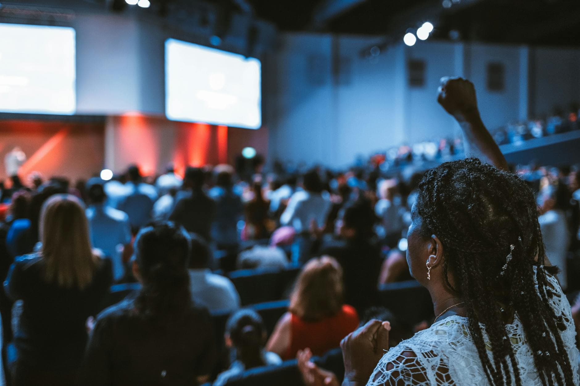 An audience member with braided hair stands and raises her hand in an engaged crowd at a dimly lit, blue-toned auditorium. The scene captures an atmosphere of collective focus or worship, with large glowing screens illuminating the stage in the distance.
