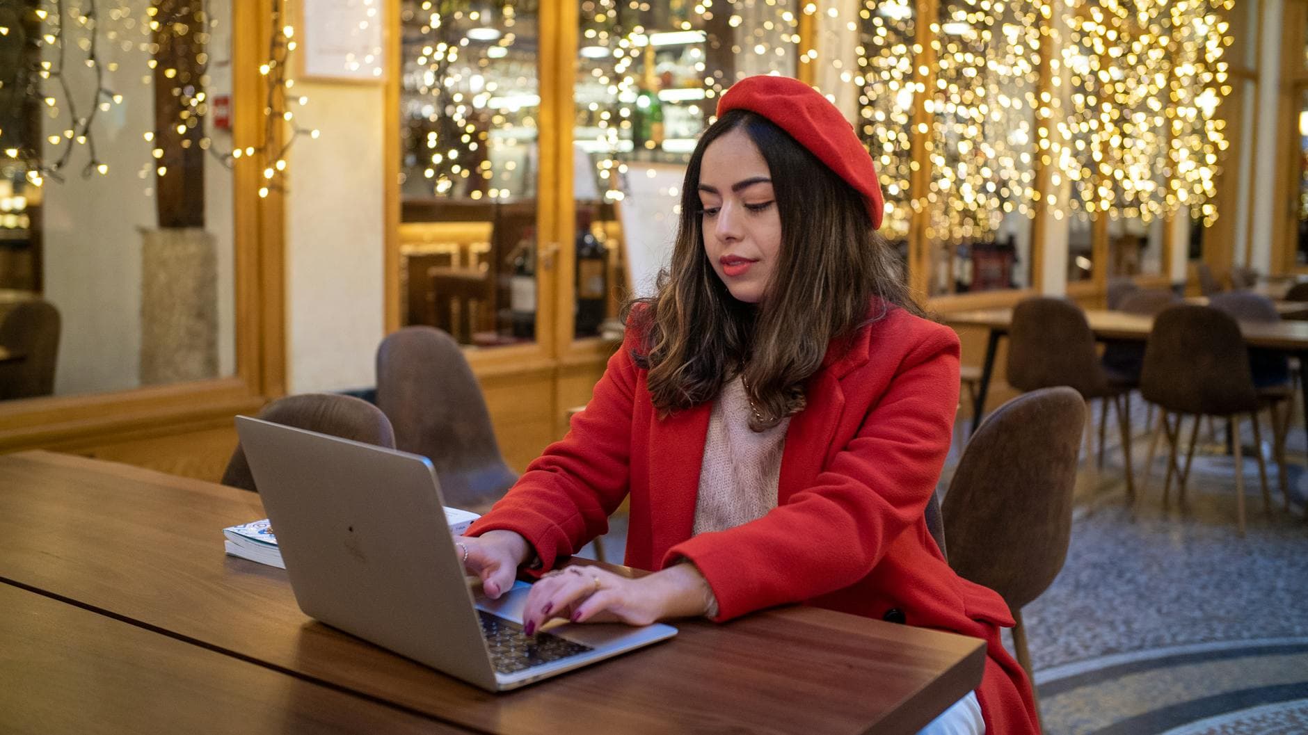 A woman in a red coat works on her laptop in a beautifully lit Parisian cafe.