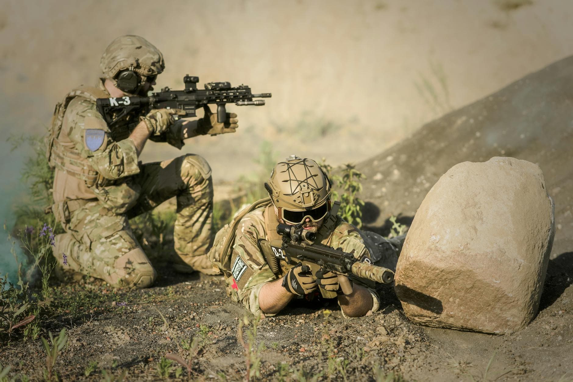 Two soldiers in camouflage uniforms practice tactical maneuvers in a desert environment, exhibiting teamwork and precision.